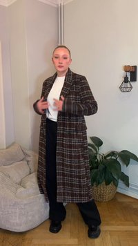 Długi brązowy płaszcz w kratę o wyraźnie zarysowanym kroju, zestawiony z białą bluzką i czarnymi spodniami. Widać fakturowaną tkaninę oraz wyraźne kieszenie.