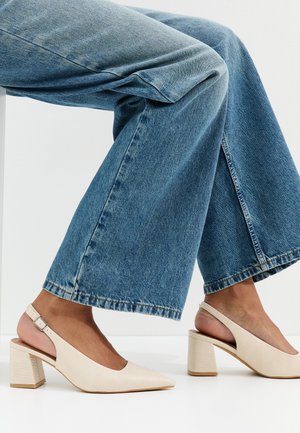 Kobieta w jasnobeżowych sandałach na obcasie typu blok na pasku za piętą, w połączeniu z niebieskimi szerokimi spodniami, siedzi na tle białego tła.