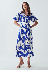 Balto audinio marškininė su drąsiais mėlynais abstrakčiais lapų raštais, trumpomis pūkuotomis rankovėmis, juosmeniu susirišamu diržu ir kulkšnies ilgio hemu.