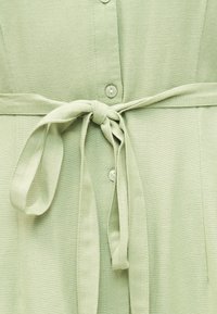 Φόρεμα από ανοιχτό πράσινο ύφασμα με κουμπιά μπροστά και ζώνη δεμένη στη μέση, που δείχνει υφή και λεπτομέρειες ραφής.