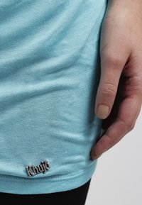 T-shirt in cotone azzurro chiaro con una texture liscia, dotata di un logo argento "Khujo" sull'orlo. Dettaglio ravvicinato della maglietta e della mano.