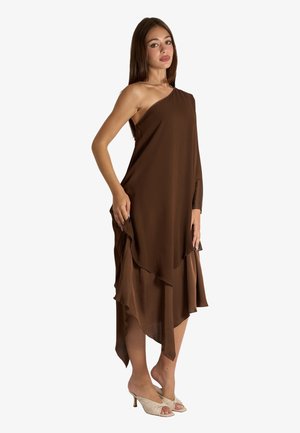 Donna che indossa un vestito marrone asimmetrico monospalla con orlo arricciato e tacchi aperti beige, in piedi davanti a uno sfondo bianco.