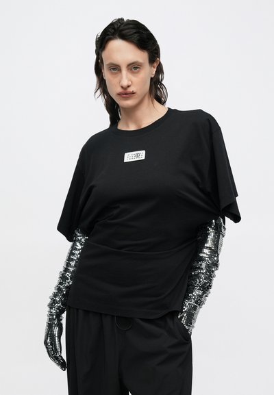 Fekete, túlméretezett póló kerek nyakkivágással, fehér címkével. Az ujjakat fémes, flitteres díszítések ékesítik. Fekete, széles szárú nadrág.