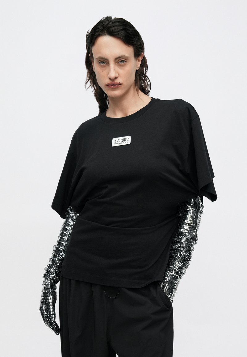 Sort oversized t-shirt med rund halsudskæring, med et hvidt mærke. Ærmer pyntet med metalliske, paliet-udsnit. Sorte bukser med vide ben.