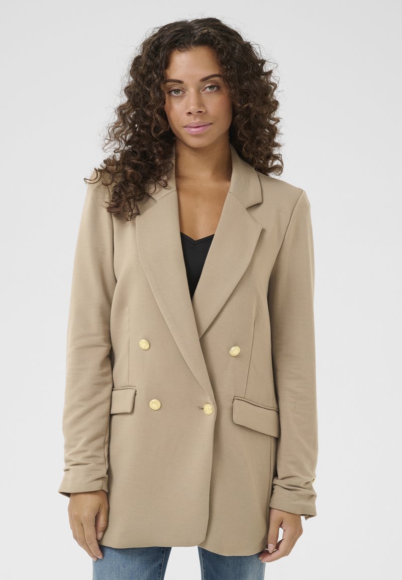 Beige dvouřadý blazer se zlatými knoflíky, klopami a předními kapsami. Hladká tkanina, dlouhé rukávy a přiléhavý střih.