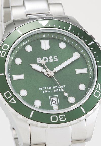 Montre en acier inoxydable avec un cadran vert, des index horaires blancs et une lunette verte assortie. Comprend un indicateur de date et une résistance à l'eau jusqu'à 50 m.