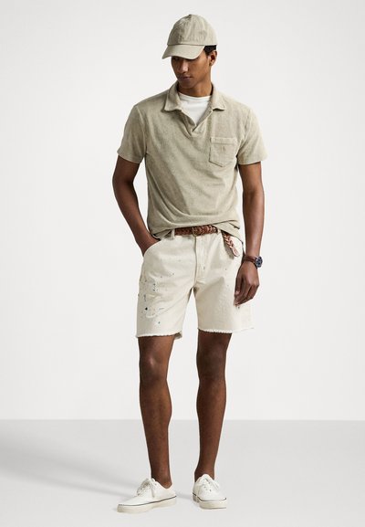 Polo shirt en velours beige avec col et poche, associé à un short beige clair avec des éclaboussures de peinture. Des baskets blanches et une casquette complètent le look.