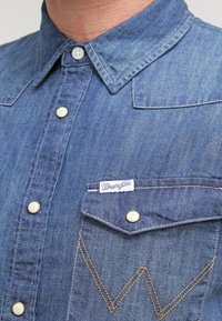 Denimskjorta i mellanblått med krage, knapptryckknappar och en bröstficka med en Wrangler-tagg och kontrasterande guldtråd i W-form.