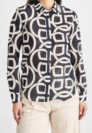 Femme portant une chemise à manches longues boutonnée avec un motif géométrique noir et blanc et un pantalon beige, debout avec les bras détendus.