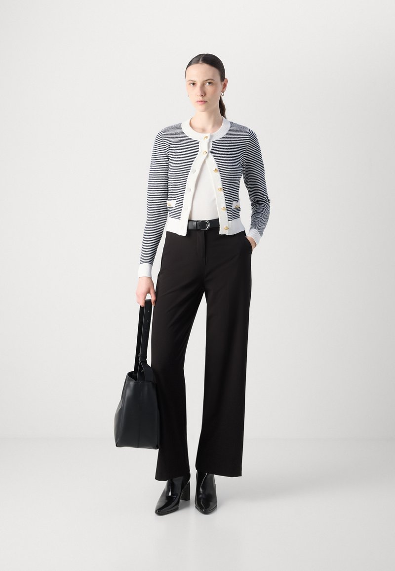Gilet tricoté avec des rayures bleu marine et blanches, boutons dorés, associé à un pantalon noir taille haute et des bottes à talons noires, tenant un sac noir.