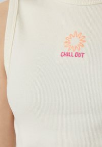 Witte geribde tanktop met een geborduurd zonontwerp in oranje en roze, met daaronder de tekst "CHILL OUT".