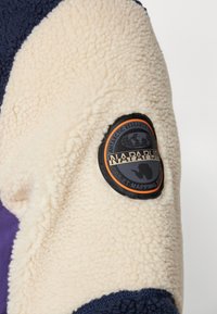 Fleece jacket with cream, navy, and purple sections. Features a circular black and orange patch labeled "Napapijri Antarctic Research."