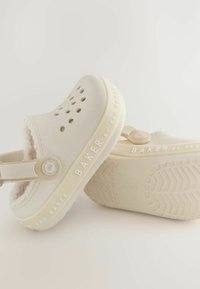 Sabots couleur crème avec des trous de ventilation, doublure douce, sangle arrière réglable et semelle extérieure texturée. Logo "BAKER par Ted Baker" sur le côté.