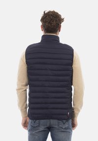 Gilet imbottito blu navy con trapuntatura verticale, colletto alto e tasche laterali con zip, indossato sopra un maglione beige chiaro e jeans blu.