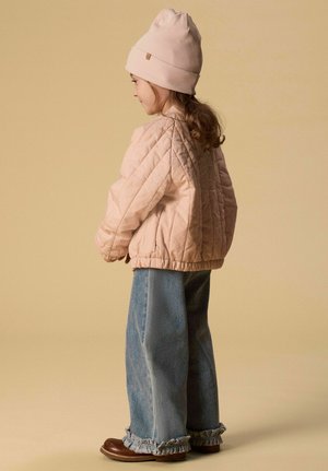 Noor tüdruk seisab külje pealt, kandes heleroosat tikitud jopet, sobivat mütsi, laia säärega siniseid teksapükse volantrist äärtega ja pruune kingi.