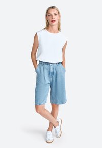 Hvid ærmeløs top, lyseblå denimshorts med plisseringer og hvide sneakers med beige såler. Enkel, afslappet outfit.