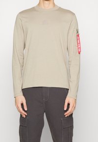 Beige pitkähihaine shirtti, joka on valmistettu puuvillasta. Rinnassa on pieni logo-patch ja hihoissa punainen vetoketju-yksityiskohta.