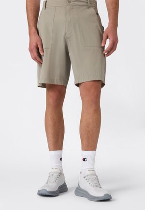 Beige Cargo-Shorts mit zwei Vordertaschen, aus leichtem Stoff. Kombiniert mit weißen Sportsocken und grauen Sportschuhen.