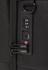 Valigia nera con superficie testurizzata, dotata di doppi zip, serratura TSA e due etichette nere con logo. Design compatto e rettangolare.