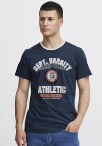 Námornícka modrá bavlnená tričko s bielym výstrihom. Na prednej strane je biely a oranžový grafický text: "Dept. Varsity Athletic Original Supply."