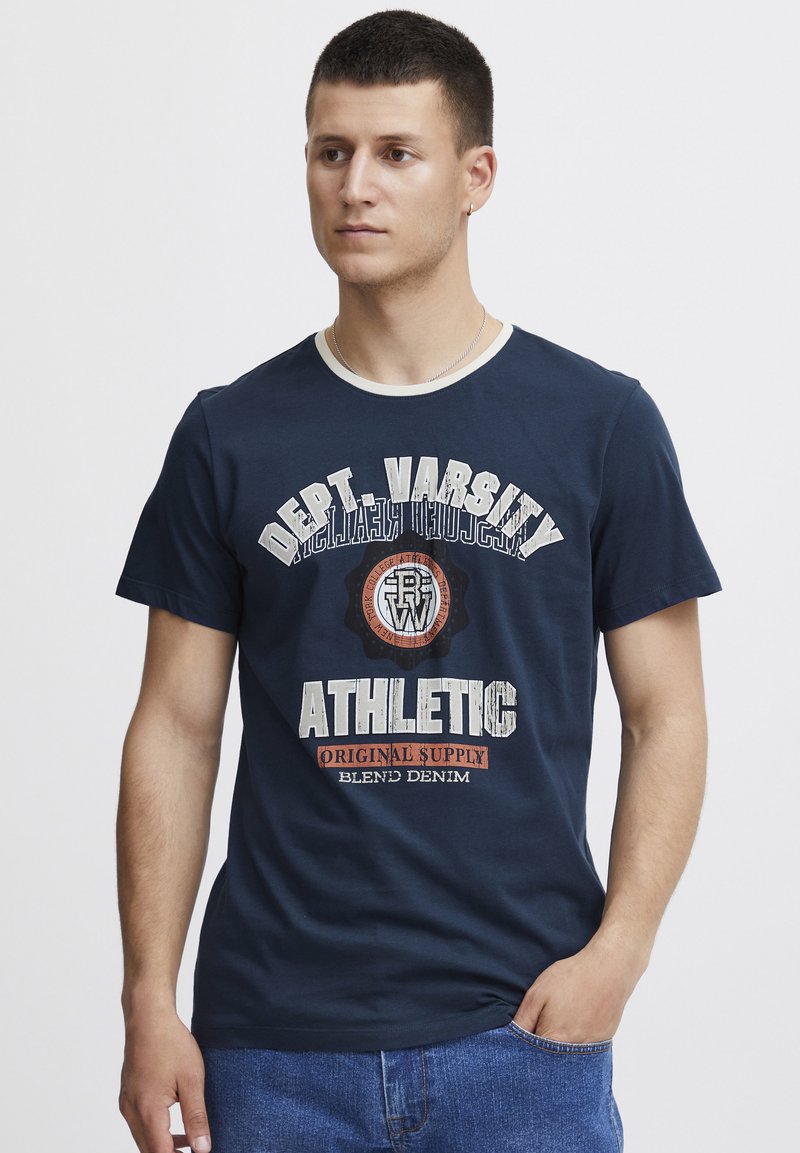 Námornícka modrá bavlnená tričko s bielym výstrihom. Na prednej strane je biely a oranžový grafický text: "Dept. Varsity Athletic Original Supply."