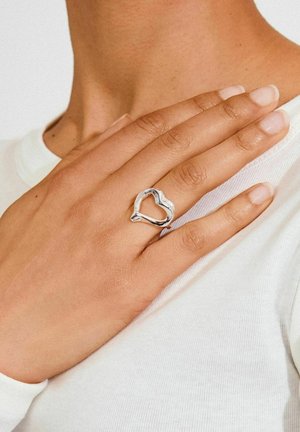 Srebrn prsten u obliku srca s uvijenim dizajnom, ističući glatke, reflektirajuće površine. Nošen na ruci s prirodnim noktima.