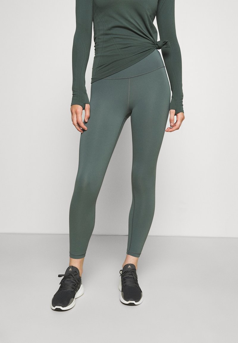 Chemise verte à manches longues avec un détail noué à la taille, associée à un legging assorti à la cheville et des chaussures de sport noires.