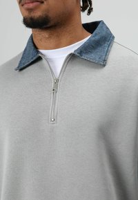 Grijze sweatjas met een spijkerbloeskragen en een zilveren rits bij de hals. Heeft een gladde textuur en een contrasterende kraagdetail.