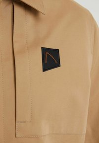 Chemise beige avec un col plié et un patch noir orné d'un motif orange. Texture lisse avec des détails de couture précis.