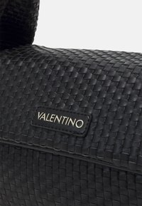 Mustasta kudotusta nahka käsilaukusta, joka on suorakulmainen muoto ja näkyvä logo-patch. Tekstuuri on monimutkainen ja pinta on matta.