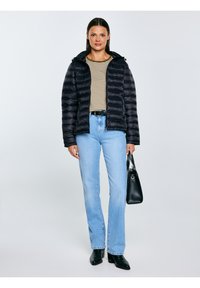 Sieviete melnā pūkainā jakā, svītrainā krekliņā, gaiši zilās džinsās, melnos zābakos un turot melnu somu uz vienkārša fona.