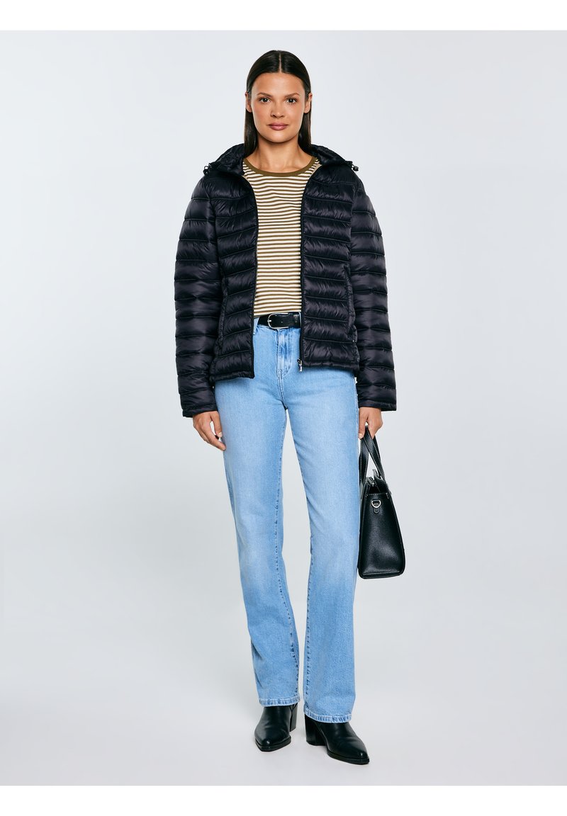 Sieviete melnā pūkainā jakā, svītrainā krekliņā, gaiši zilās džinsās, melnos zābakos un turot melnu somu uz vienkārša fona.