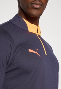 Sötétkék, negyedcipzáros sportfelső narancssárga gallérral és Puma logóval. Textúrált anyag, testhezálló fazon, sportoláshoz ideális.