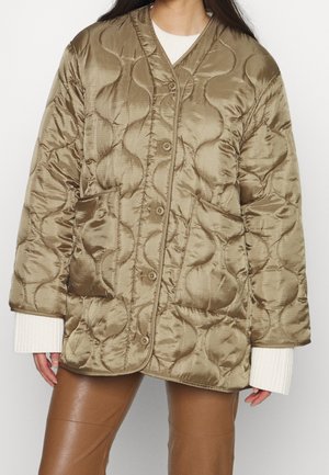 Frau trägt übergroße, gesteppte olivgrüne Jacke mit großen Vorderseiten-Taschen und Knopfverschluss, kombiniert mit brauner Hose und weißen Pulloverärmelbündchen.