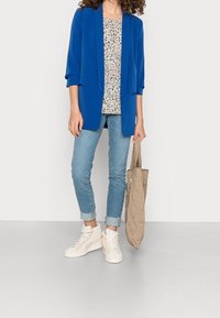 Blazer ajusté bleu vif, haut léger à motifs, jean délavé clair, sac cabas beige matelassé, et baskets crème. Tissu lisse, coupe décontractée.