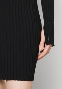 Robe noire côtelée à manches longues, avec une coupe ajustée et une fente au niveau du poignet. Le tissu a une texture douce et un léger éclat.