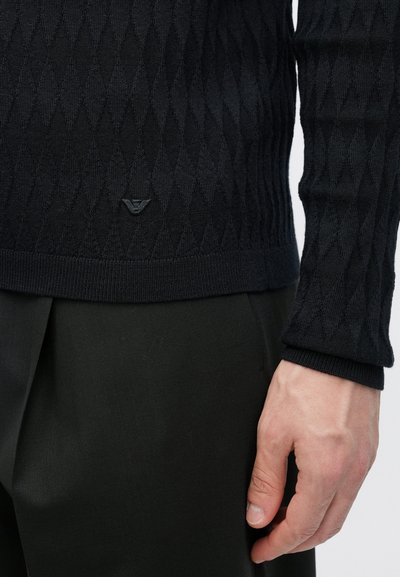 Μαύρο πλεκτό πουλόβερ με υφή διαμαντένιου σχεδίου, με ένα μικρό λογότυπο κοντά στο τελείωμα, σε συνδυασμό με σκούρο παντελόνι που φαίνεται στο πλάι.