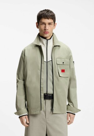 Ung mand iført en lysegrøn lynlåstrøje over en hvid højhalset sweater og beige ternede bukser mod en ensfarvet hvid baggrund.