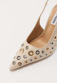 Beige leather high-heeled shoe with a pointed toe, adorned with metallic circular eyelets in gold, silver, and black. Smooth texture.