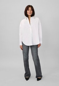 White long-sleeve button-up shirt with a relaxed fit, curved hem, paired with gray flared jeans and black pointed-toe heels.