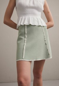 Zelená pletená mini sukně s bílými scalloped okraji a svislým bílým prošíváním. V kombinaci s bílým smockovaným topem s volánovým lemem.