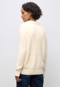 Maglione beige chiaro con una finitura morbida e testurizzata, dotato di colletto sciallato e maniche lunghe, indossato sopra jeans in denim scuro.