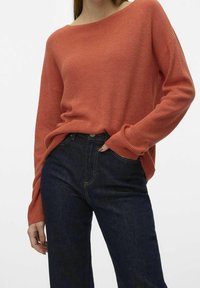 Femme portant un pull en tricot loose couleur rouille, légèrement rentré dans un jean bleu foncé taille haute, avec une main dans la poche.