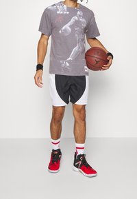 Grå T-shirt med en basketball grafik, sort og hvid shorts, røde basketballsko og armbånd, holder en basketball.