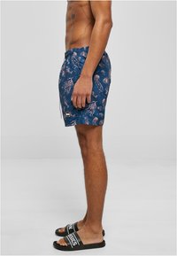 Bañador azul con estampado de medusas rosas, cinturilla elástica y cordón. Chanclas negras con texto blanco.