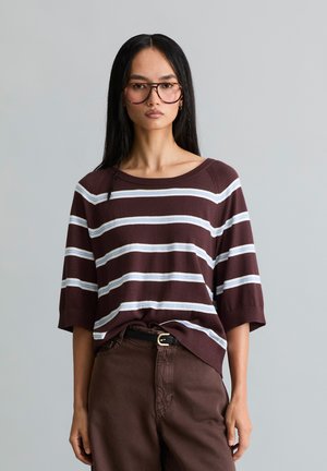 Femme aux longs cheveux foncés et lunettes, portant une chemise marron à rayures à manches courtes rentrée dans un pantalon marron foncé, sur un fond uni.