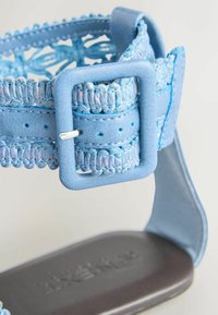 Gros plan d'une bride de sandale bleu clair avec une boucle carrée texturée et des détails tissés sur une semelle grise.