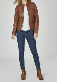 Giacca in pelle marrone con tasche con cerniera, abbinata a una maglietta bianca e jeans skinny blu scuro, completata con stivali alla caviglia marroni.