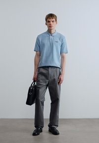 Ανοιχτό μπλε πόλο πουκάμισο με οριζόντιες ρίγες, σε συνδυασμό με γκρι παντελόνια και μαύρα δερμάτινα παπούτσια. Κρατάει μια μαύρη δερμάτινη τσάντα.