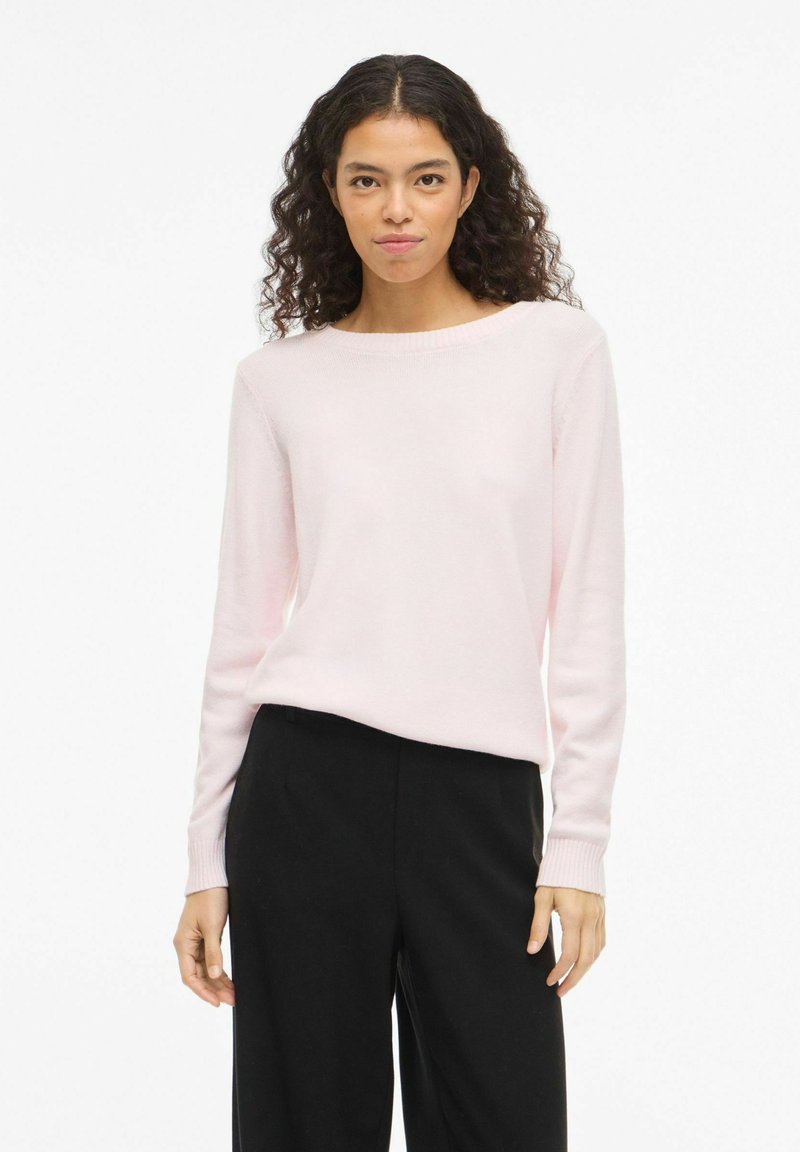 Femme aux cheveux bouclés et foncés, portant un pull à manches longues rose clair et un pantalon noir, debout devant un fond blanc uni.